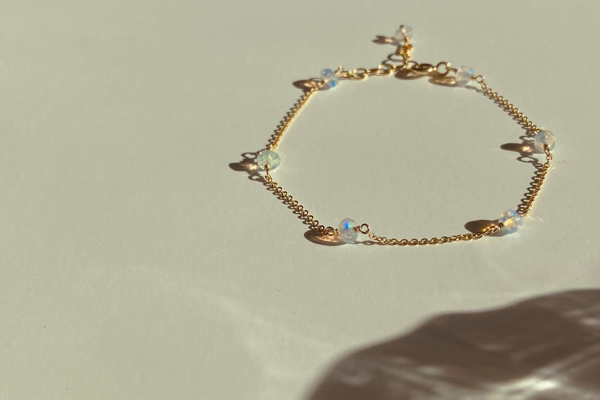 A delicate gold opal bracelet with small opal beads, photographed on a warm off-white surface with a soft shadow.