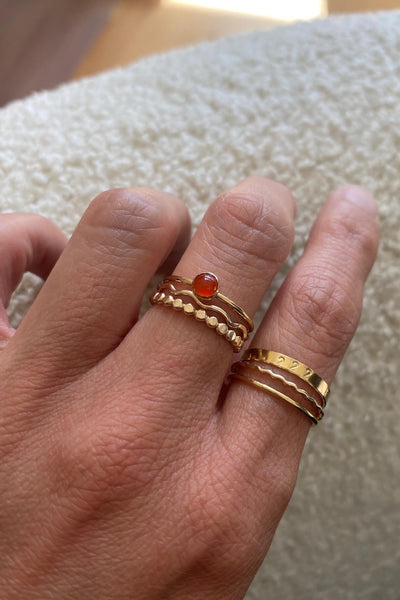 A hand wearing gold stacking rings including a carnelian ring, wave ring and hammered texture dot ring in the middle finger and index finger wearing a 222 ring, wave ring and a thin stackable gold ring.